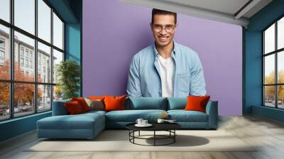 Smiling handsome young man with cheerful expression, has toothy smile, wears transparent spectacles, wears denim shirt, rejoices good work, isolated over purple background. Positive emotions Wall mural