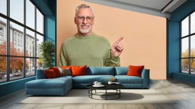 Smiling positive senior man with grey hair and beard points right and shows perfect copy space has brilliant smile white teeth wears optical glasses casual jumper poses against beige background Wall mural