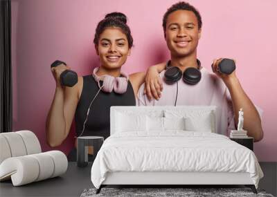 Sporty motivated woman and man lift dumbbells for training muscles do exercises in gym dressed in sportswear use headphones for listening music isolated over pink background. Healthy lifestyle concept Wall mural