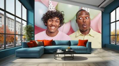 Studio photo of young cheerful smiling broadly Hindu and surprised African american men standing close to each other among colorful balloons celebrating birthday having party spending good time Wall mural