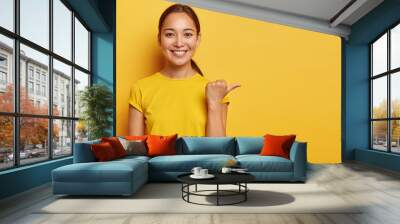 Studio portrait of young cheerful Asian female points away with thumb, happy face expression, demonstrates copy space for advertisement, has pleasant appearance, wears bright yellow clothes. Wall mural