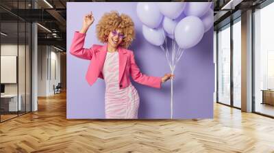 Studio shot of cheerful carefree lady enjoys special occasion organises party holds bunch of inflated balloons wears frestive long dress pink jacket and sunglasses poses against purple background Wall mural