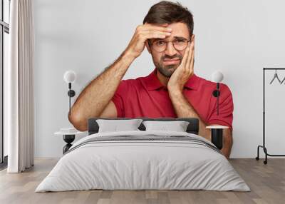 Studio shot of dissatisfied bearded male has toothache and headache, feels unhappy and tired after long work, frowns face in discontent, dressed in red t shirt, isolated over studio background Wall mural