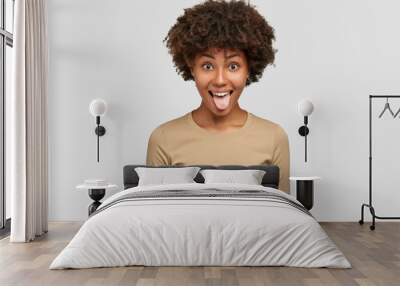 Studio shot of funny black woman shows tongue, has playful expression, Afro hairstyle, makes grimace, dressed in casual beige sweater, isolated over white background. Comic dark skinned girl Wall mural