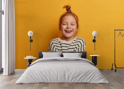 Studio shot of ginger child embraces herself, expresses self love, closes eyes from pleasure, wears striped jumper, poses against yellow studio wall, feels good, shows white teeth, full of excitement Wall mural