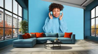 Studio shot of good looking woman with curly hair wears stereo electronics headphones enjoys listening music podcast wears warm sweater isolated over blue background. Pleased female meloman. Wall mural