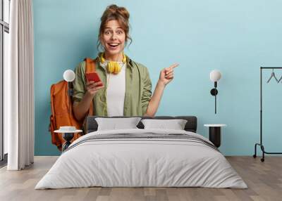 Studio shot of happy teenage girl holds mobile phone, checks email, gets new notification, uses modern headphones, points at copy space for brand name or label, wears loose khaki shirt, carries bag Wall mural