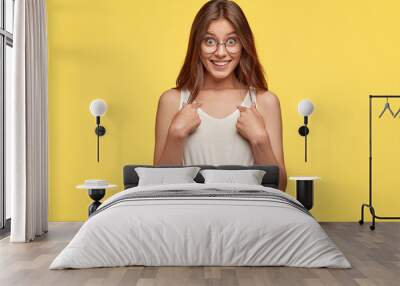 Studio shot of indignant woman with pleasant appearance, points at herself as asks she really won prize, has cheerful expression, dressed in casual vest and shirt around waist, isolated on yellow Wall mural