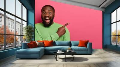 Studio shot of overjoyed bearded Afro American man points at blank space, keeps mouth opened, wears green jumper, isolated over pink background, shares cool link, asks advice and picks promo Wall mural