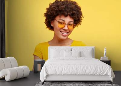 Studio shot of pleasant looking ethnic woman with gentle smile, expresses gratitude, has arms folded on chest, wears t shirt and shades, isolated over yellow studio wall. Truthful feelings from heart Wall mural