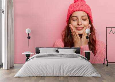 Studio shot of pleasant looking female model touches chin with both palms, has eyes closed, enjoys delicate well cared skin, has dark hair, dressed in winter outfit, isolated on pink wall, blank space Wall mural