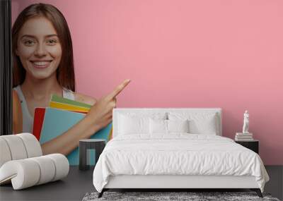 Studio shot of positive Caucasian woman with shining smile, dark straight hair, poins at upper right corner, shows free space for your advertisement, ready for school, isolated over pink background Wall mural