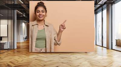 Studio shot of pretty young Indian girl points index finger at mock up copy space demonstrates new offer or great opportunities has happy smile on face wears shirt isolated over brown background Wall mural