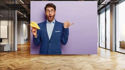 Studio shot of surprised employer or executive manager in elegant blue suit, points aside with thumb, stupefied notice something strange, holds paper airplane in hands, isolated on purple background. Wall mural