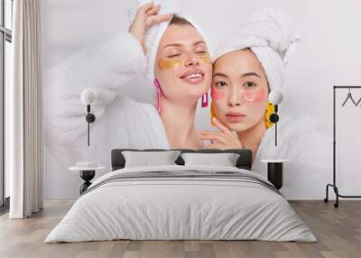 Studio shot of two mixed race young women wear moisturising beauty patches undereyes after taking shower dressed in bathrobes towels over head isolated over white background take care of skin and face Wall mural