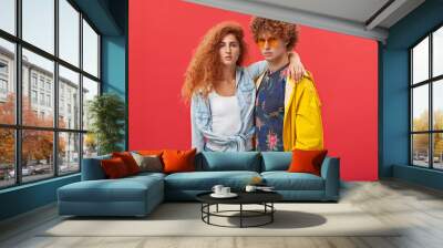 Style and fashion. Attractive confident young male and female models posing at blank studio wall, dressed in trendy clothes, looking at camera with serious facial expressions; girl hugging guy Wall mural