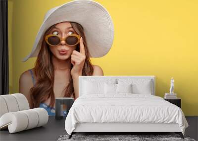 Stylish woman with surprised mysterious expression, looks asdie, wears stylish summer shades, hat and polka dot blouse, isolated over yellow background with copy space for your text. Relaxation Wall mural