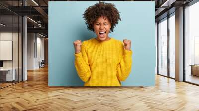 Successful dark skinned female student happy to get scholarship, clenches fists, accomplishes goal, exclaims finally victory, stands amused over blue background. Success, cheer and achievement concept Wall mural