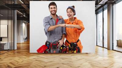 Successful ecstatic young Caucasian man and female wearing overalls and carrying working instruments bumping fists together as sign of celebration, feeling happy and excited about doing great job Wall mural