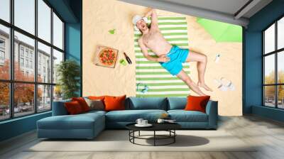 Summer holidays and recreation concept. Shocked young man lies on green striped towel at sandy beach eats delicious pizza drinks beer enjoys lazy day sunny weather wears panama and blue shorts Wall mural