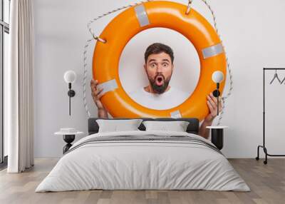 Suprised bearded man holds inflated lifebuoy stares bugged eyes wide opened mouth learns to swim ready to rescue you dressed in casual t shirt isolated over white background. Male beach rescuer Wall mural