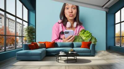 Surprised Asian woman holds mobile phone checks eaten calories in app carries fresh green vegetables keeps to healthy nutrition wears pink windbreaker and headphones around neck poses indoor Wall mural