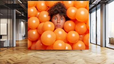 Surprised birthday girl poses over inflated orange party balloons stares bugged eyes expresses great wonder or surprise. Young Afro American woman enjoys anniversary event has shocked expression Wall mural