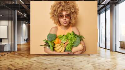 Surprised curly haired young woman keeps lips folded poses with green grocery fullof vitamins eats seasonal vegetables and fruits wears sunglasses and t shirt isolated over beige background. Wall mural