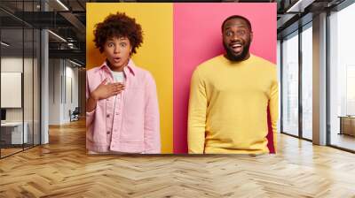 Surprised woman gasps from wonder, keeps hand on chest, happy dark skinned guy stands near, giggles positively over funny joke. Afro American couple express different emotions shock and positiveness Wall mural