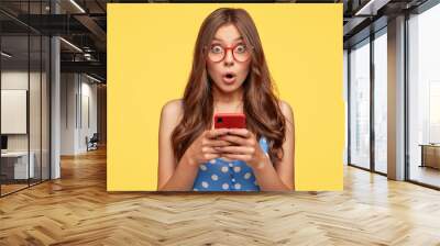 Surprised young female model holds modern cell phone, reads sudden news on internet web page, surfes in social networks, poses against yellow background. People, emotions, technology, lifestyle Wall mural