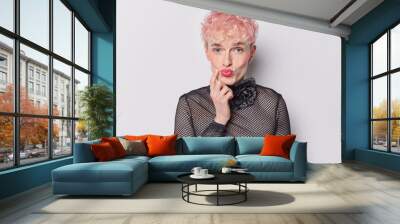 Surprised young man with pink hair keeps lips rounded has finger on cheek wears black net jumper poses against white background. Handsome transgender guy poses in studio. Human facial expressions Wall mural