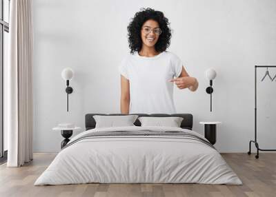 T-shirt design and advertising concept. Style and fashion. Indoor shot of cheerful smiling young African American woman with curly hair pointing index finger at copy space on her blank white top Wall mural