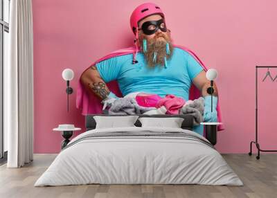 Thoughtful bearded husband in superhero costume has fun and poses near basket full of dirty laundry detergent focused aside poses against pink background. Household chores cleanliness concept Wall mural