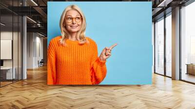 Thoughtful blonde forty years old European woman wears spectacles and knitted orange sweater points away on blank copy space shows place for your advertisement isolated over blue background. Wall mural