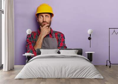 Thoughtful man builder construction touches chin, thinks over new idea for building, works as repairman, wears yellow protective helmet, checkered shirt and apron. Industry and repairing concept Wall mural