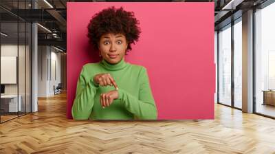 Time to go, hurry up! Shocked ethnic woman points at wrist, makes time gesture, shows we should do everything quickly, dressed in green clothes, stands against pink background. Its too late. Wall mural