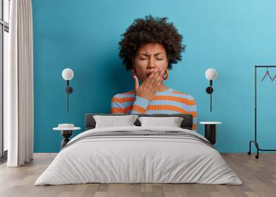 Tired exhausted Afro American woman feels bad after hard day at work, covers mouth and has sleepy expression, wears casual striped jumper, isolated on blue background. Restless and sleepiness concept Wall mural