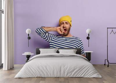 Tired overworked male student yawns, drinks coffee from red mug to feel refreshed, opens mouth, wears sailor oversized jumper, wants sleep, poses over purple background. People and drinking concept Wall mural