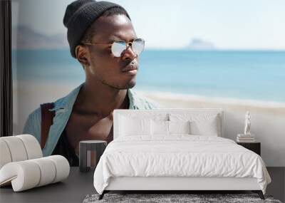 Tourism, travel, leisure, lifestyle and adventure. Attractive stylish dark-skinned traveler dressed in trendy clothes enjoying beautiful seascape during nice walk on urban beach early in the morning Wall mural