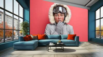 Travel adventure and winter sport concept. Puzzled frozen woman with red icy face looks embarrassed at camera trembles from cold wears ski goggles grey coat goes hiking in stormy snow weather Wall mural