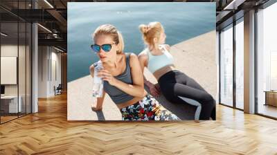 Two attractive female runners having rest after jogging exercise sitting on the beach. Two young athlete blonds resting and drinking water during physical workout outdoors on sunny morning
 Wall mural