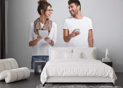 Two energetic happy young European colleagues man and woman standing at desk side by side and having conversation, discussing business issues, plans and results, using modern electronic devices Wall mural