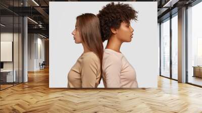 Two mixed race women stand in profile back to each other, have thoughtful expressions, dressed in casual clothes, think about cooperation, isolated over white background. Studio shot. Ethnicity Wall mural