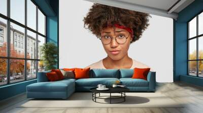 Unhappy dark skinned girl looks with displeased upset expression, frowns face, feels upset, wears glasses, casual sweater, isolated over white background. People and negative emotions concept Wall mural