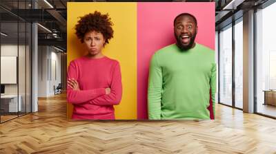 Unhappy offended woman stands with arms crossed, doesnt speak to husband. Joyful bearded man has dark skin, giggles positively at something funny, expresses positive emotions. People, reaction Wall mural