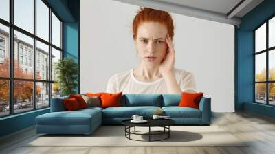 Unhappy stressed young redhead female with hair knot touching face while suffering from bad headache, frowning and looking at camera with tensed and painful expression on her face. Body language Wall mural