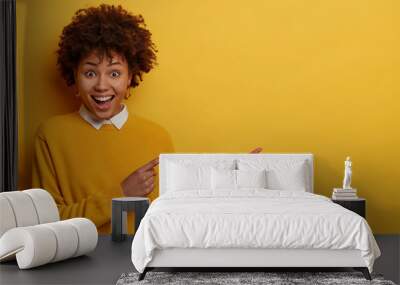 Upbeat positive young woman with Afro hair shows something awesome, attracts your attention to discounts, wears yellow clothes, gestures indoor, involved in advertising campaign, promots item Wall mural