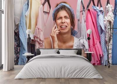 Upset woman standing near rack of clothes, chatting over smart phone with her friend, complaining that she has nothing to wear. Displeased female not knowing what to put on for birthday party Wall mural