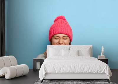 Vertical image of joyful young girl feels overjoyed, raises clenched fists, being in good mood, wears white sweater and pink hat, dressed in warm clothes during cold autumn day, isolated on blue wall Wall mural