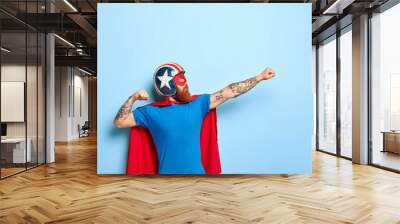 Vertical shot of bearded man makes flying gesture, clenches fists, has goal to achieve, wears protective headgear, red cape, pretends being heroic character, has superhuman power isolated on blue wall Wall mural